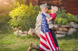A soldier in uniform kneeling and embracing a young child, who is holding an American flag, set in a green outdoor environment with plants and rocks.