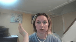 A woman with medium-length hair and makeup is sitting in a room, speaking and gesturing with her right hand. She is wearing a gray shirt and a pearl necklace, with a staircase and artwork visible in the background.