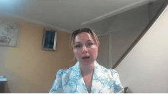 A woman wearing a light-colored floral blouse speaks while seated in a room with a staircase in the background.