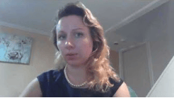 A woman with wavy hair wearing a pearl necklace, seated in a room with a light-colored wall and an abstract painting in the background.