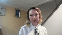 A woman with medium-length blonde hair, wearing a white shirt with a collar and a bow tie, is speaking in a well-lit room with a staircase in the background.