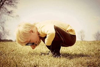 A young child with blonde hair crouches on a grassy field, examining something closely with a magnifying glass.