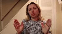 A woman in a patterned blouse is using hand gestures while speaking, with a staircase in the background.