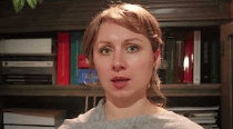 A woman with light brown hair and a gray sweater, facing the camera in front of a bookshelf filled with books.