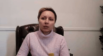 A woman sitting in a leather chair, wearing a light pink sweater, speaking directly to the camera.