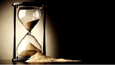 An hourglass with sand flowing from the upper chamber to the lower chamber, set against a dark background.
