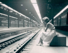 A woman in a white dress sitting on a suitcase at an empty train station, deep in thought, with tracks and overhead lights visible.