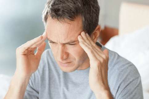 A man sitting on a bed with a pained expression, holding his head as if experiencing a headache or stress.