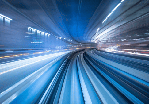 A high-speed photographic capture of a moving train or transit system, with blurred blue and white lights creating a dynamic sense of motion along curved tracks.