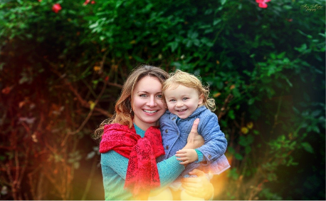 A smiling woman holding a young girl outdoors with a lush green background and flowers.