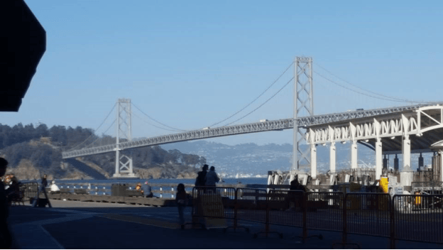 A view of a suspension bridge spanning over water, with people walking on a path in the foreground and hills in the background.