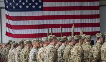 A group of soldiers in military uniforms standing in formation, with a large American flag displayed in the background.
