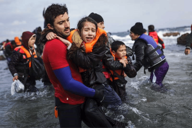 A man in a red shirt carries a young girl in a life jacket as they wade through water, surrounded by other individuals wearing life jackets. The scene captures a moment of rescue amidst a turbulent sea.