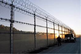 A security fence topped with barbed wire, with a person standing near a vehicle at sunset.