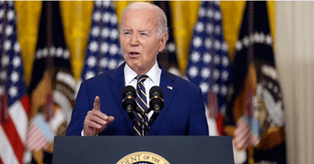 President Joe Biden delivering a speech at a podium with the Presidential seal, surrounded by flags.